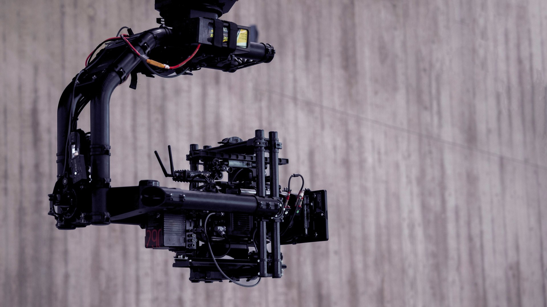 Foto einer Film-Kamera vor einer grauen Wand. Die Kamera kommt von oben und hängt an einem Stativ.