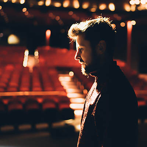 Michael Jener Profilbild. Michael Jener steht vor leeren Stühlen in einem leeren Theater. Der Hintergrund ist unscharf.