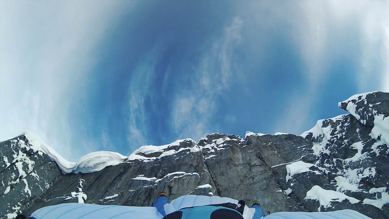 Eine Person in einem Wingsuit springt einen Berg runter. Man sieht nur den Rücken und die Beine der Person.