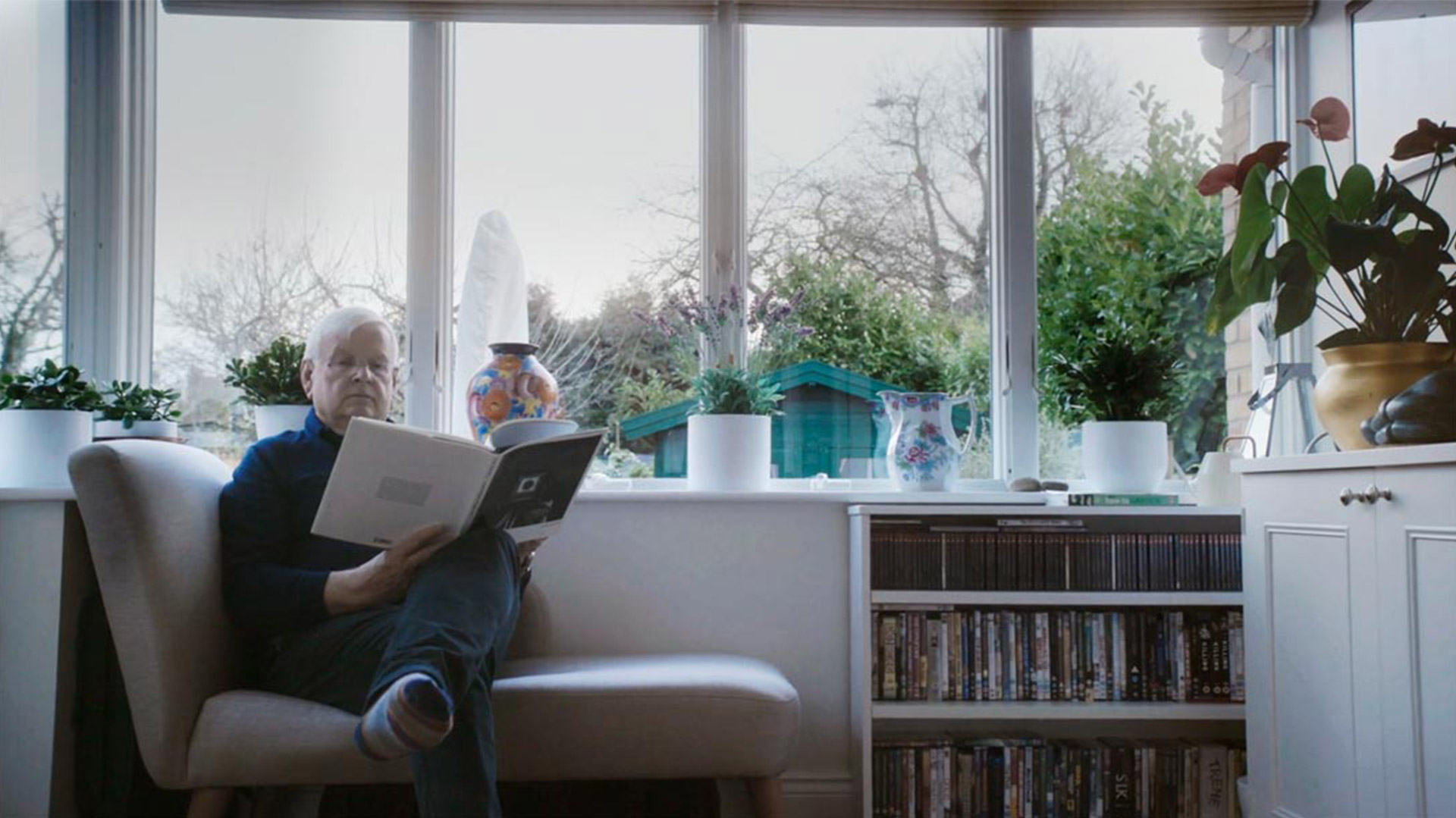 Bild eines Zimmers, auf der linken Seite sitzt ein Mann mit einem Buch auf einem Sessel. Links ist ein Regal mit CD's und Filmen. Im Hintergrund sieht man durch Fenster einen Garten.