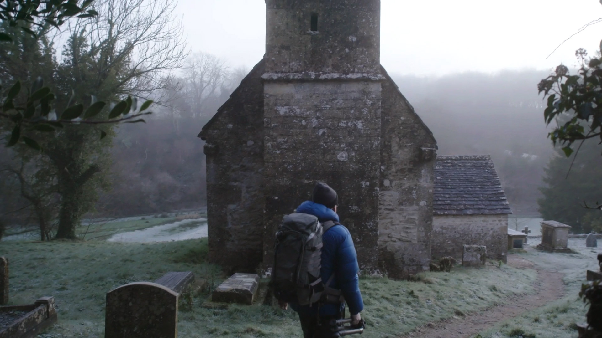 Ein Mann geht auf eine ältere Kapelle zu, in der Hand hält er ein Stativ und er trägt einen großen Rucksack.