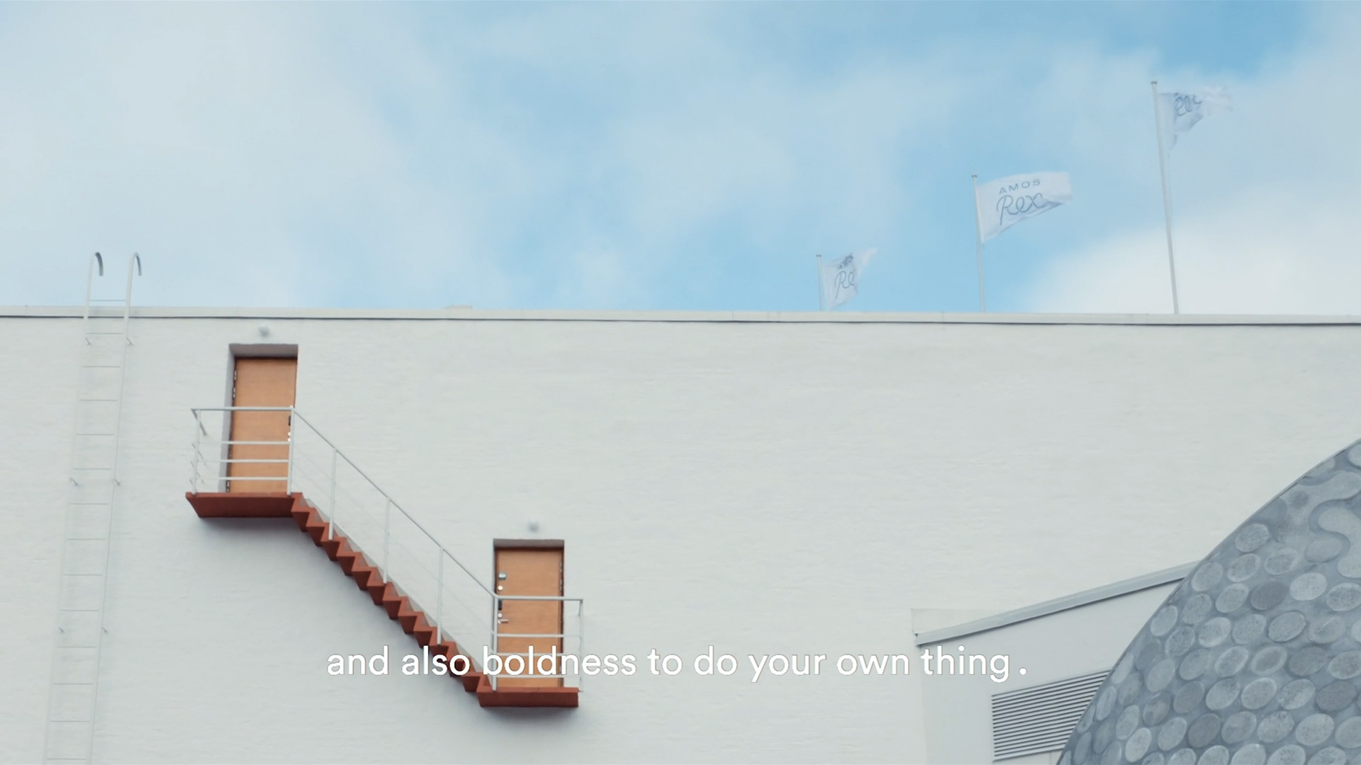 Es ist eine weiße Mauer oder Wand zu sehen, oben sieht man den Himmel. An der Wand sind zwei Türen die durch eine Treppe verbunden sind.